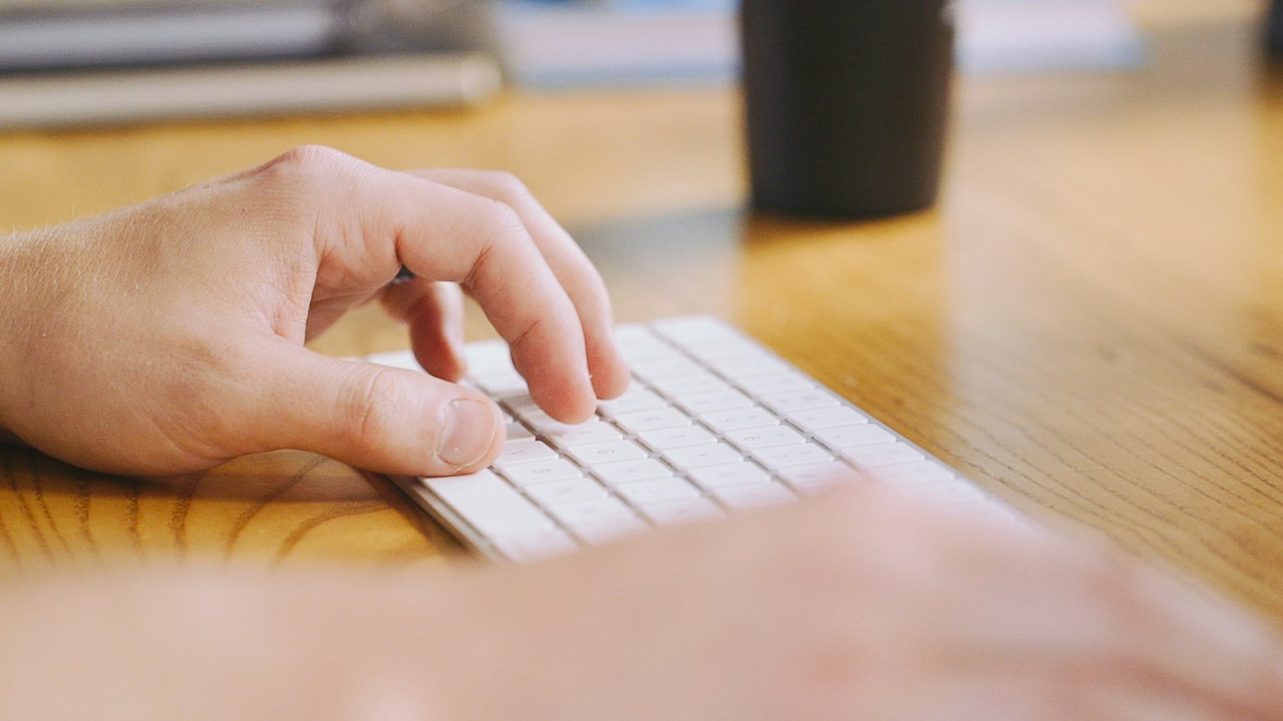 Manos escribiendo en un teclado blanco sobre un escritorio de madera.